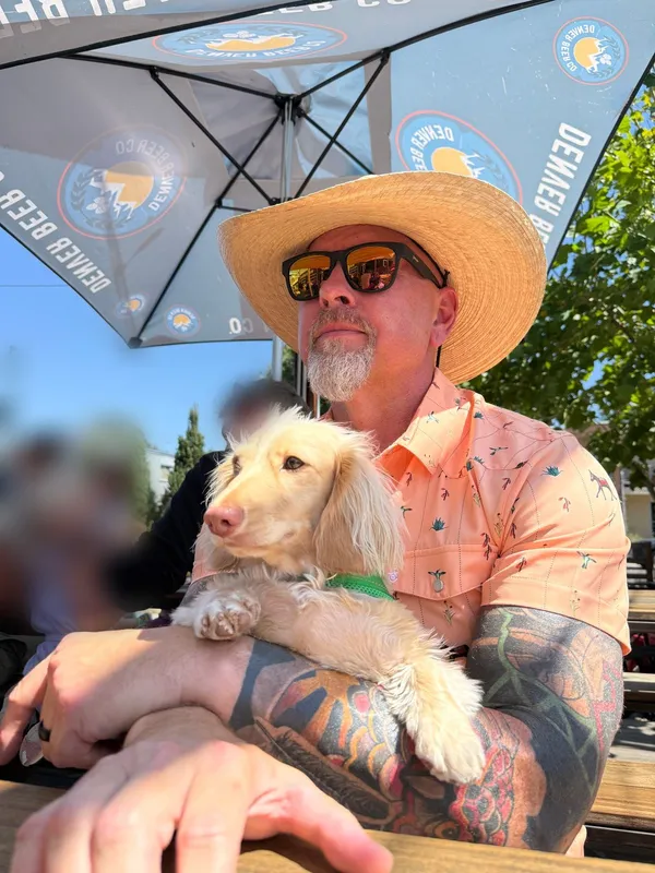Richard Bird in a straw hat and mirrored sunglasses at an outdoor table, holding a cream long-haired dachshund