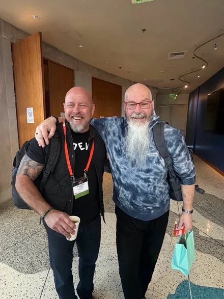 Richard Bird and Chris Roberts in a conference hallway, arms round each other's shoulders, both laughing — Bird in a black t-shirt and lanyard holding a coffee, Roberts in a tie-dye hoodie with a long white beard and red glasses
