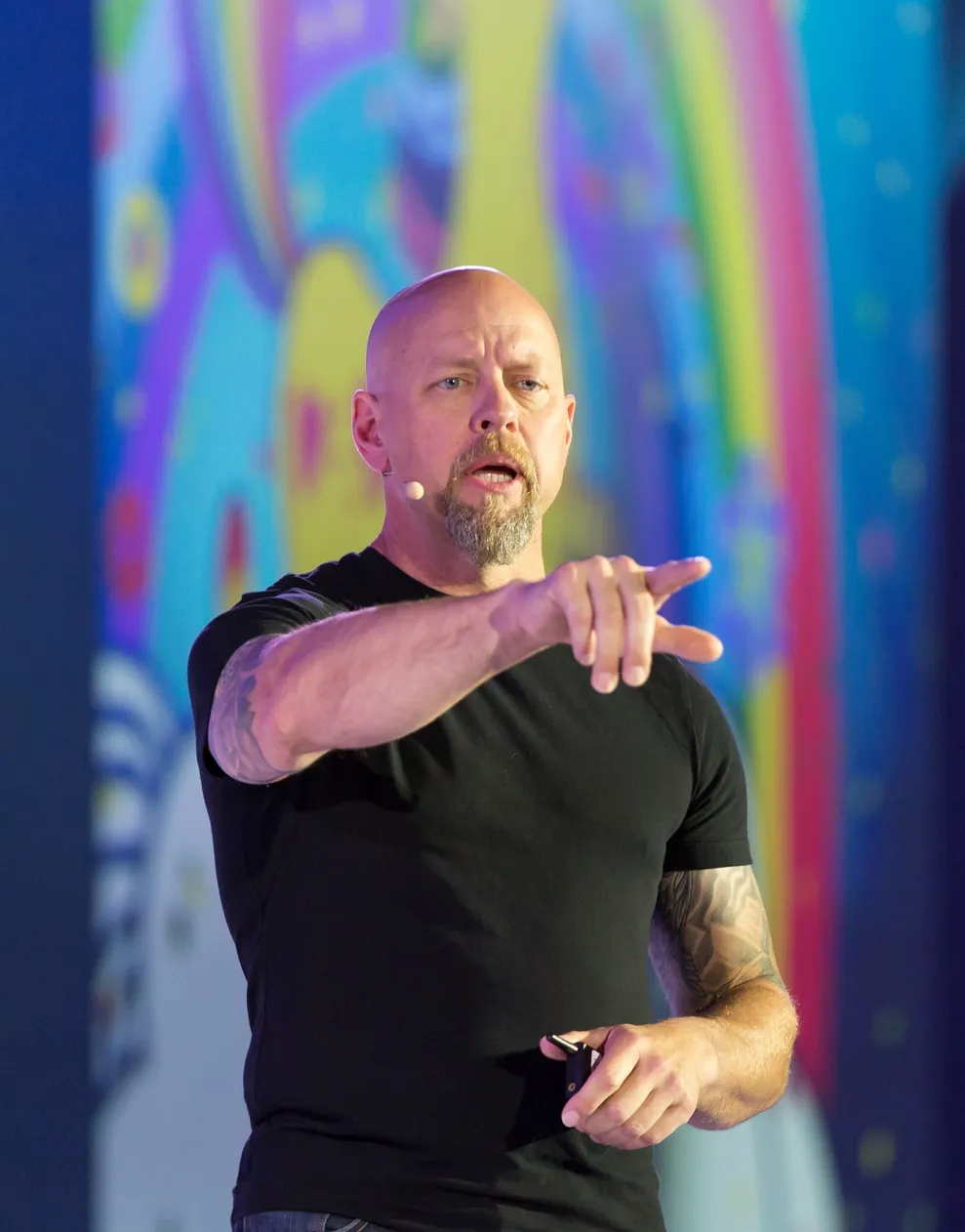 Richard Bird on stage mid-sentence in a black t-shirt, tattooed forearm, pointing out into the audience, a large screen of saturated color behind him
