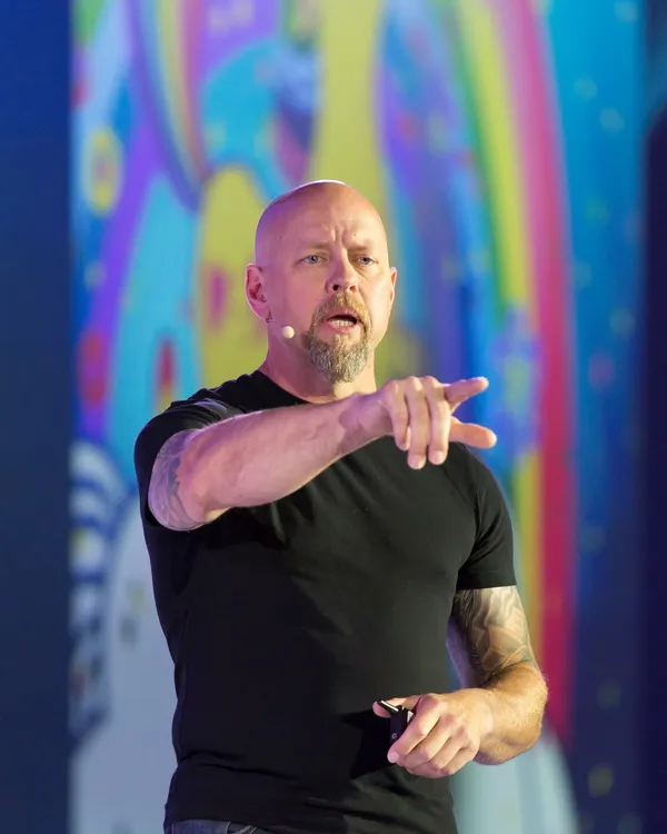 Richard Bird on stage mid-sentence in a black t-shirt, tattooed forearm, pointing out into the audience, a large screen of saturated color behind him