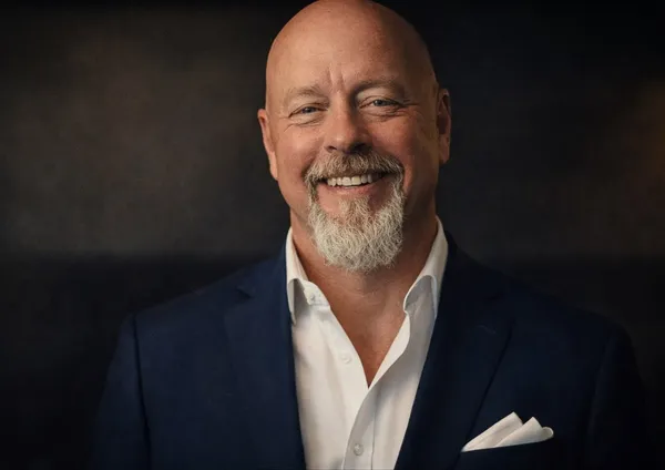 Studio headshot of Richard Bird smiling, in a navy jacket over an open-collared white shirt with a white pocket square, against a dark backdrop