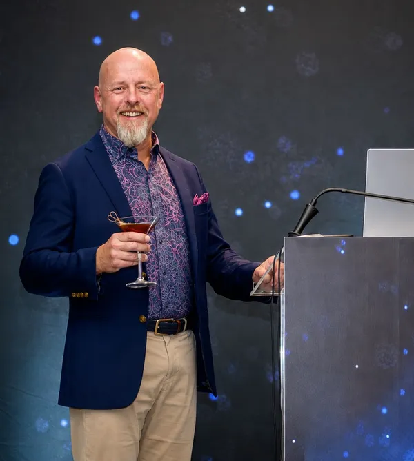 Richard Bird standing at a lectern in a navy blazer and patterned shirt, raising a Manhattan, against a dark backdrop scattered with blue points of light