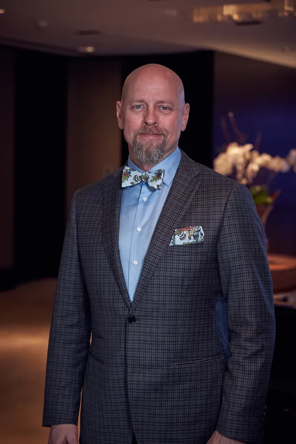 Richard Bird standing in a gray check suit, pale blue shirt, floral bow tie and matching pocket square, in a warmly lit interior